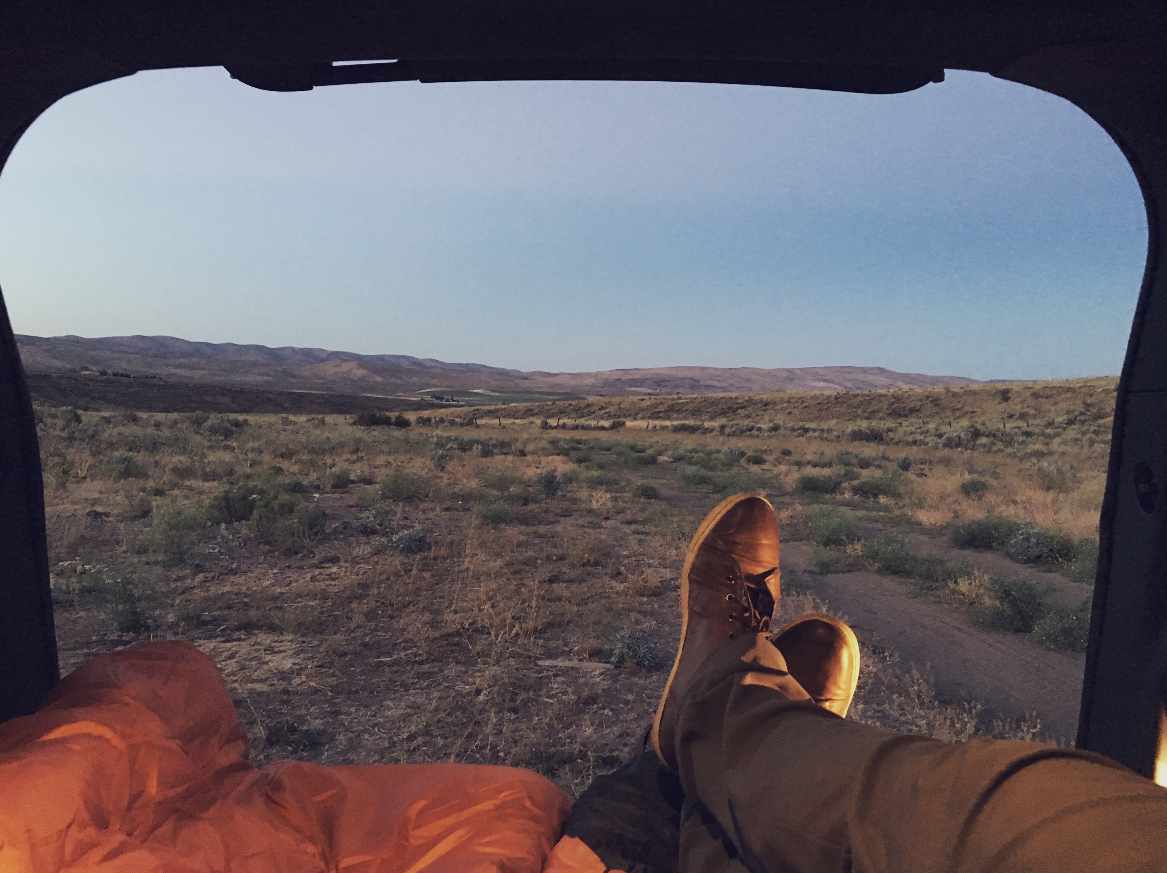 Feet propped out of a truck window with a sleeping bag, looking out at a high desert landscape at dawn