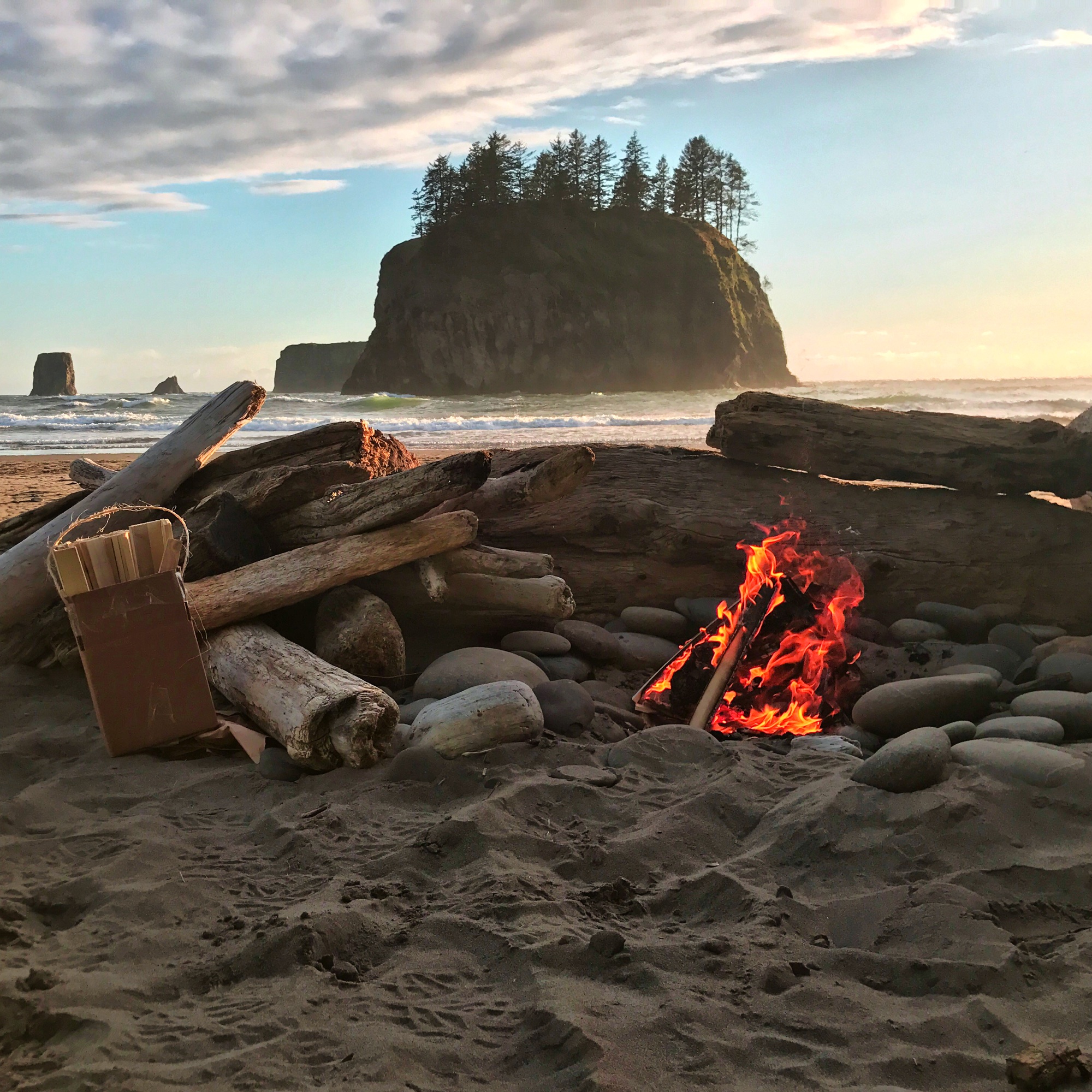 Campfire burning on a Pacific Northwest beach with dramatic sea stacks in the background