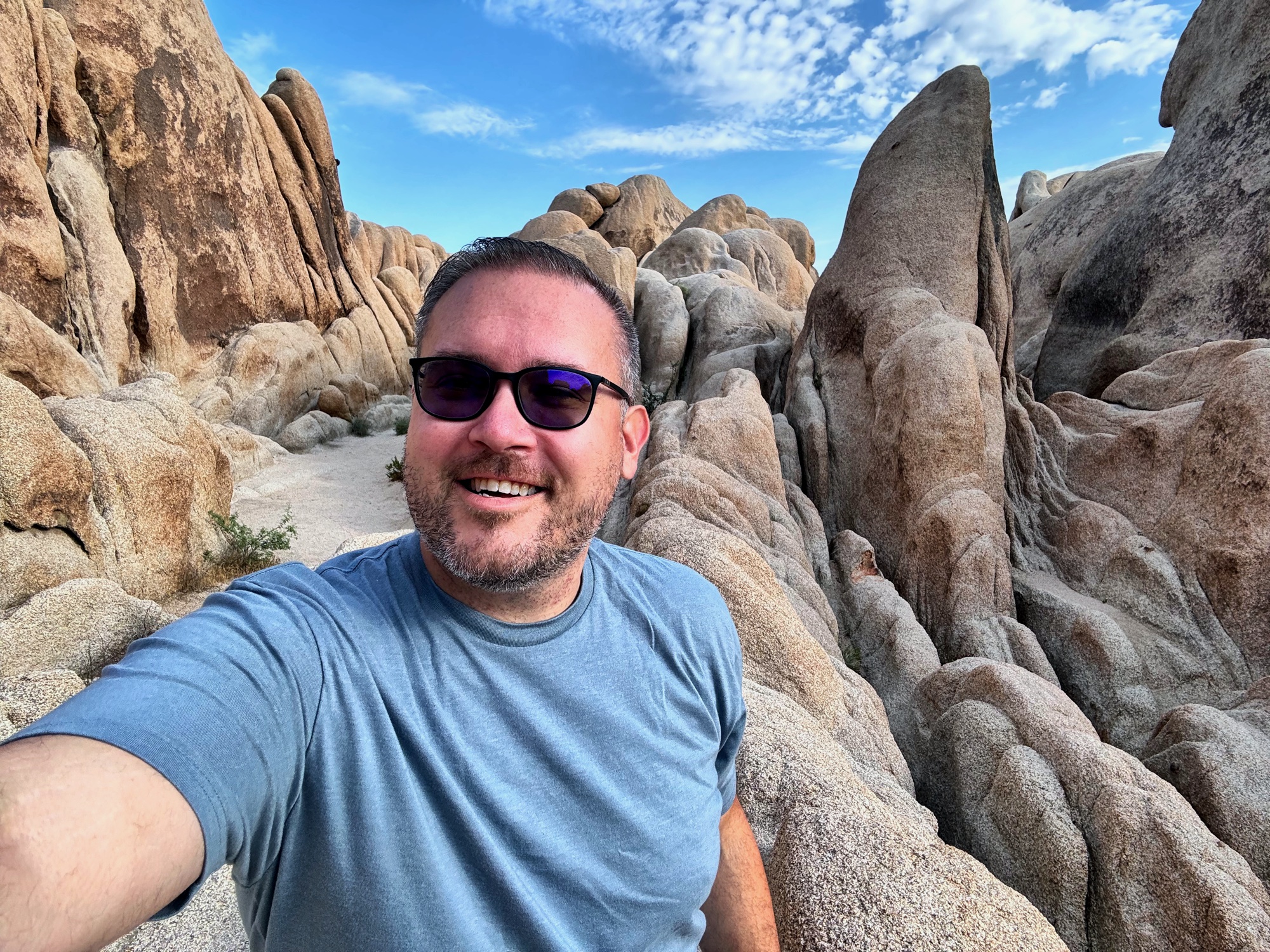 Jon grinning in a selfie surrounded by massive smooth granite boulders under a bright blue desert sky