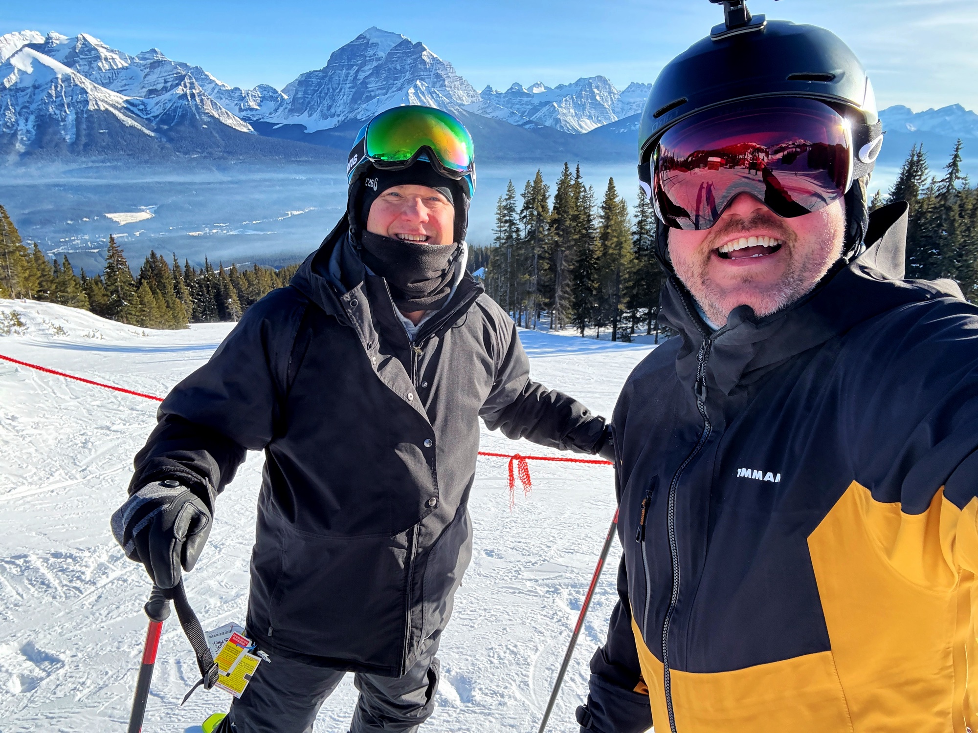 Jon and a friend smiling on skis with the dramatic snow-capped peaks of the Canadian Rockies stretching behind them