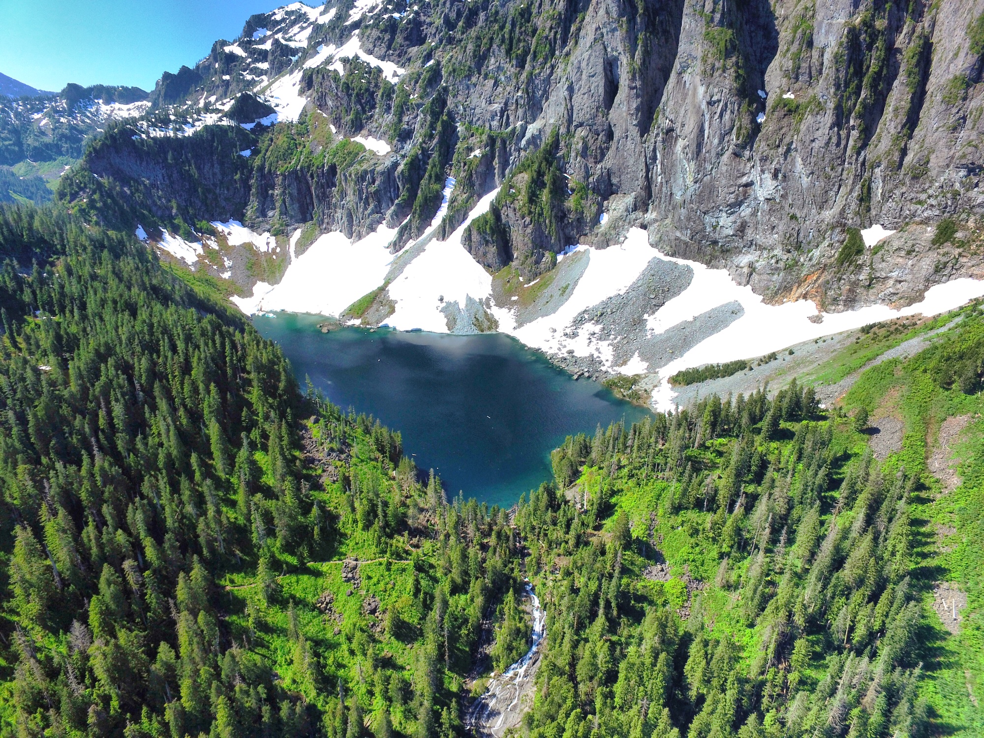 Aerial view of a deep blue alpine lake nestled in a steep valley surrounded by snow patches and dense evergreen forest