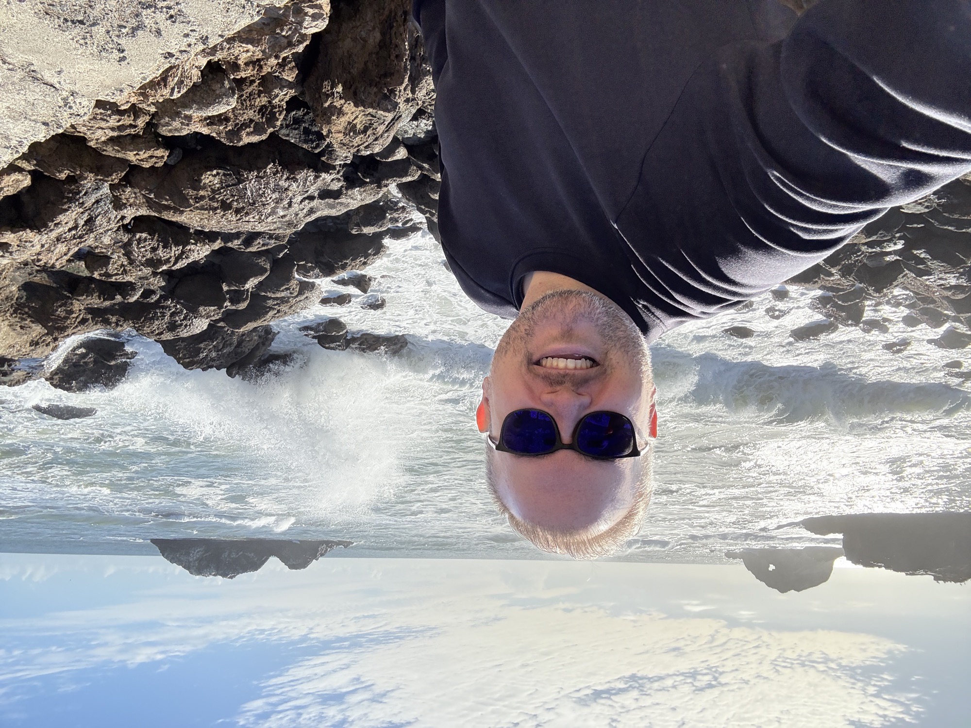 Jon smiling on a rocky coastline as a wave crashes against jagged rocks and sea stacks rise out of the ocean behind him