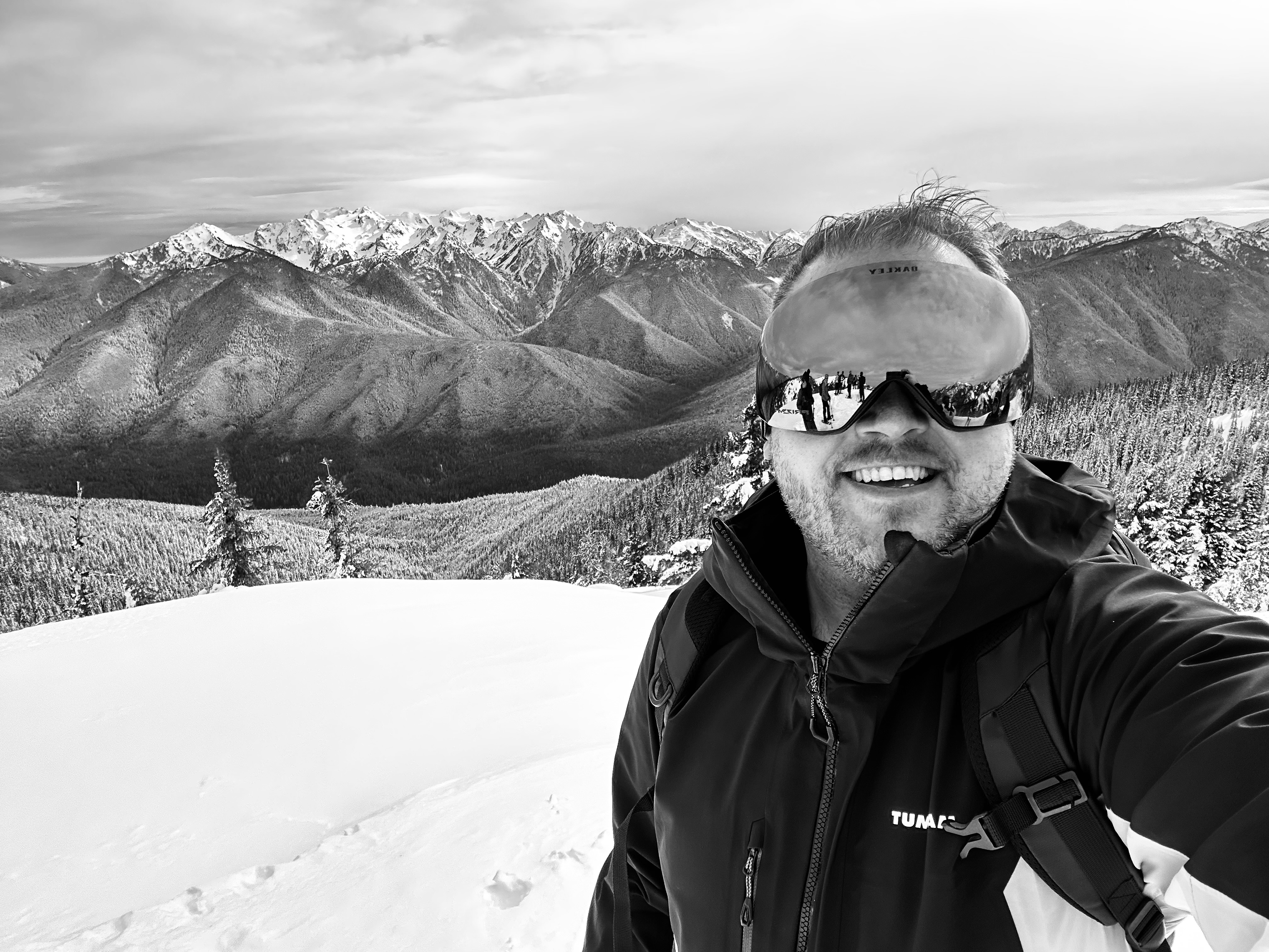 Jon grinning in a black and white selfie with ski goggles on his head and a dramatic snow-covered mountain range stretching behind him
