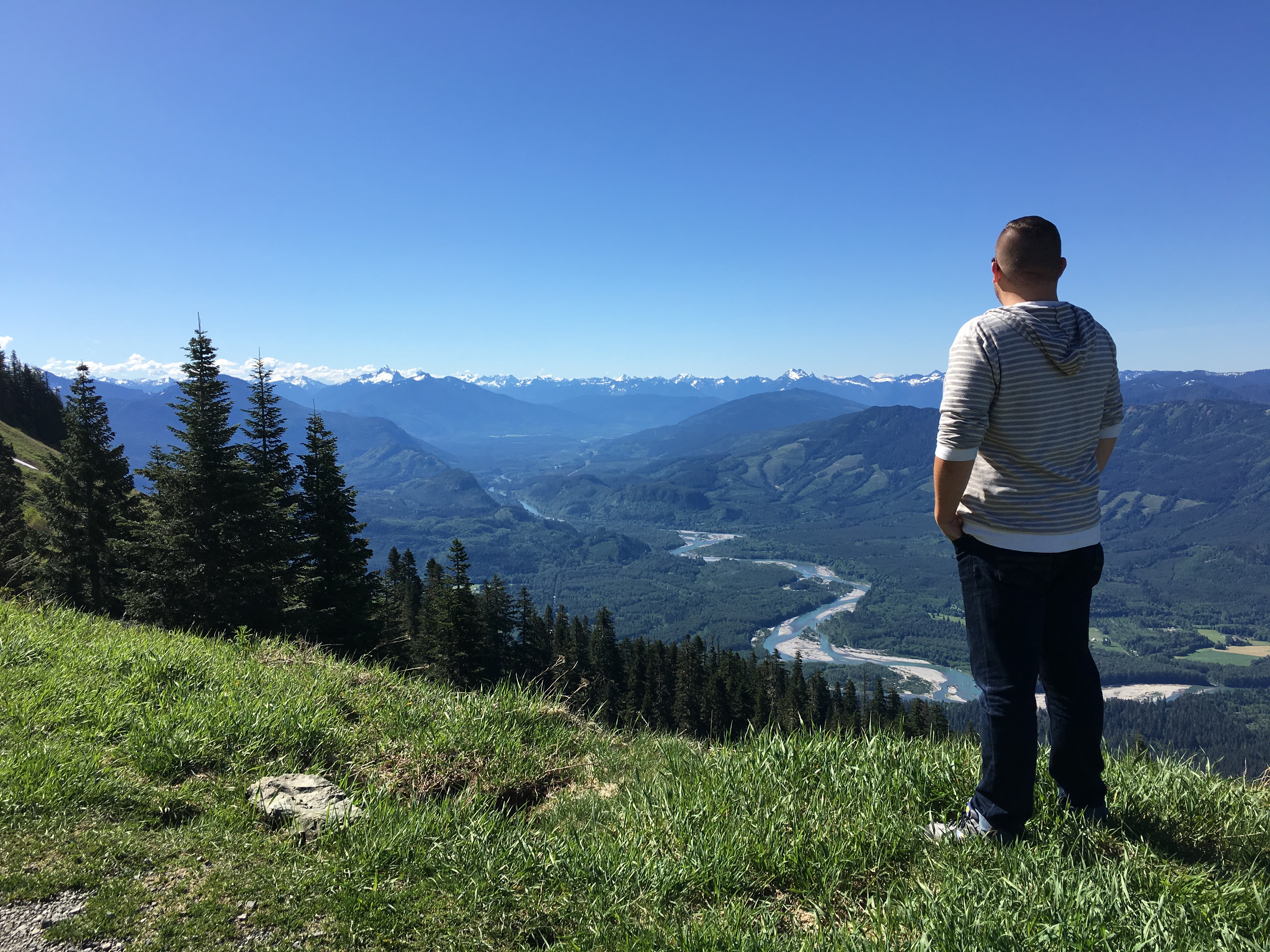 Jon standing with his back to the camera looking out over a wide river valley with snow-capped mountains on the horizon under a deep blue sky