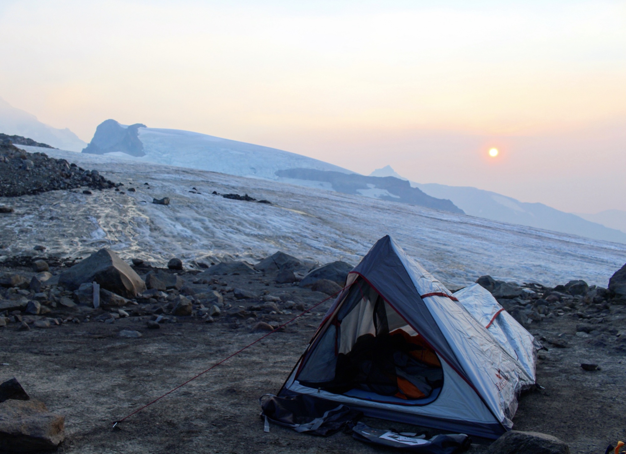 A small tent pitched on a rocky glacier moraine at sunset, sun glowing orange through smoky haze over the ice