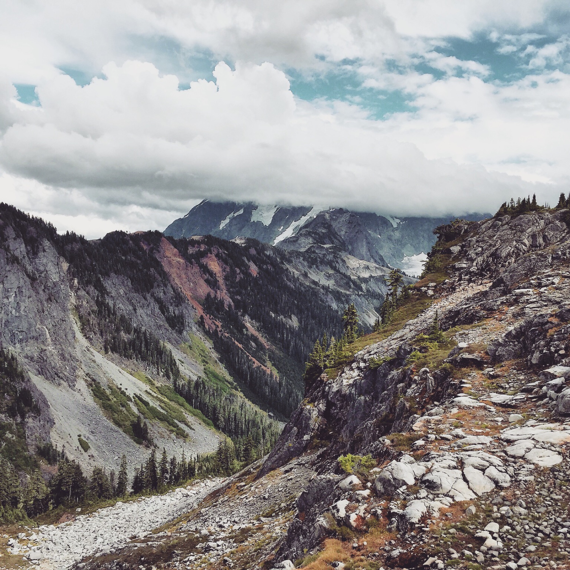 Rocky alpine trail winding through a dramatic mountain valley with moody clouds over snow-streaked peaks