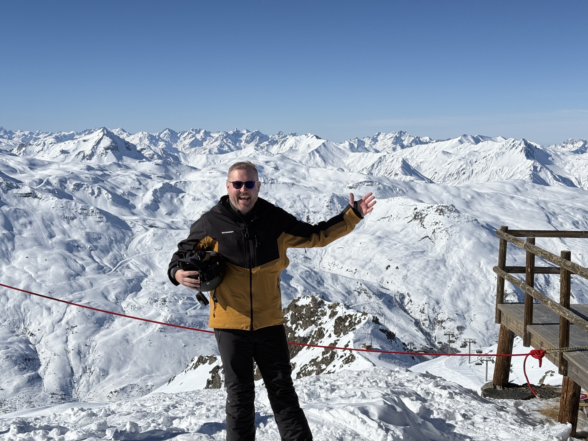 Jon standing at a ski summit with an enormous panorama of snow-covered Alps stretching endlessly behind him under a deep blue sky