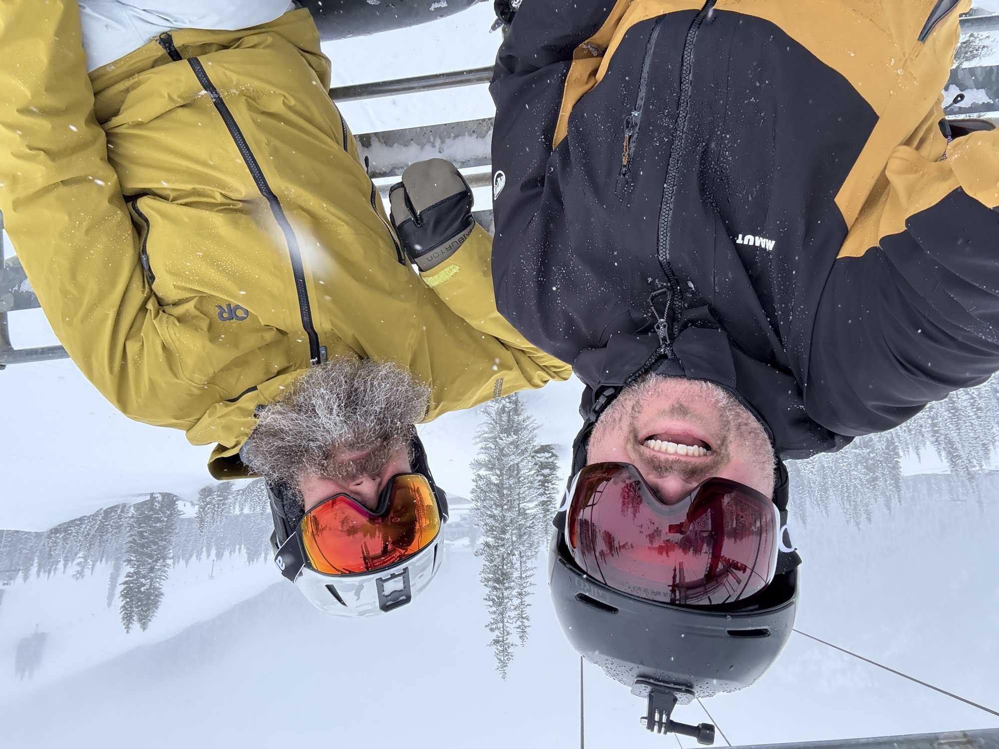 Jon and a bearded friend on a chairlift, snow falling hard, both grinning behind ski goggles in a whiteout storm