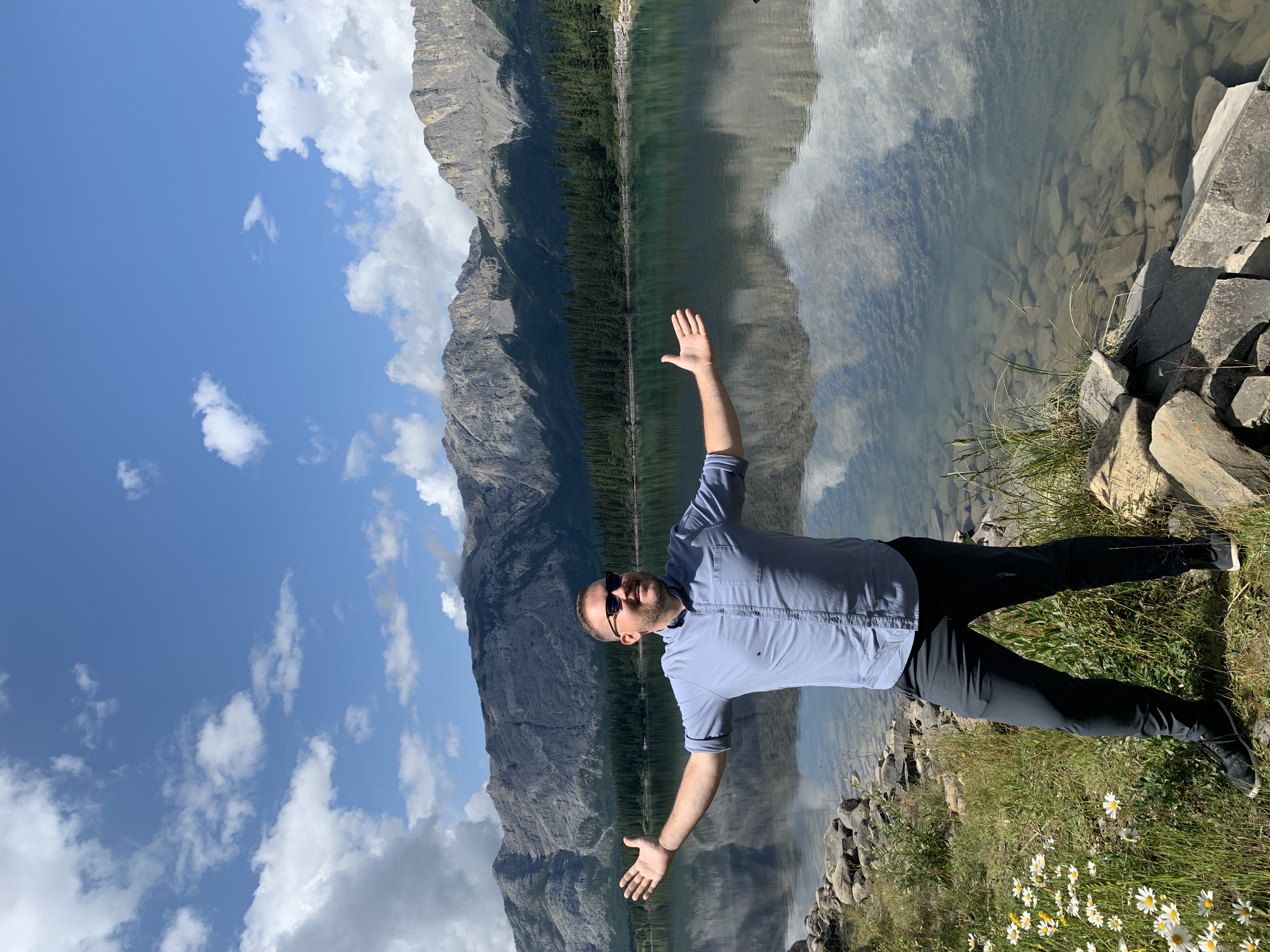 Jon posing with arms out next to a perfectly still glacial lake with mountains and sky reflected in the water, wildflowers in the foreground