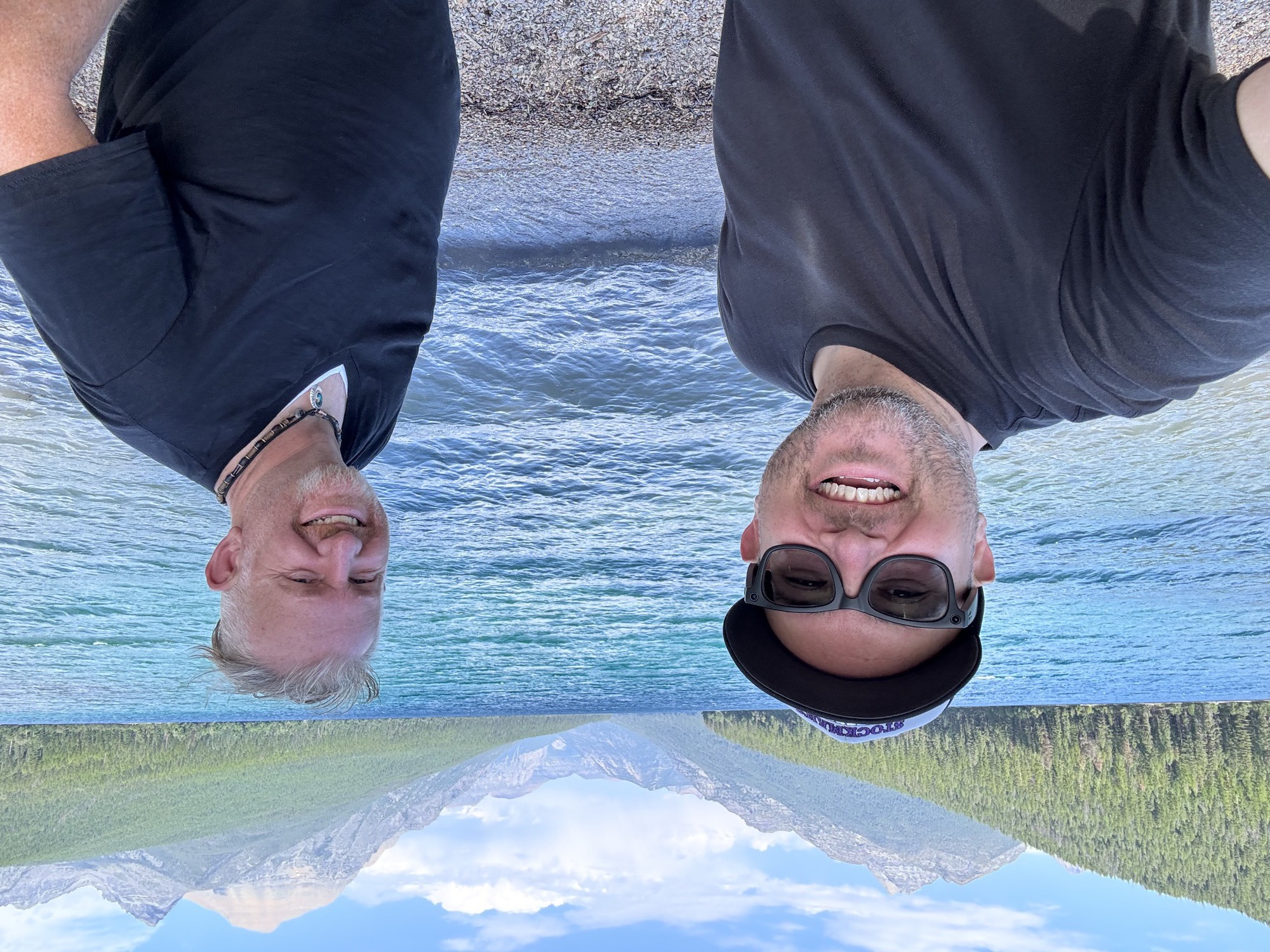 Jon and a friend smiling in front of a brilliant turquoise mountain lake with forested peaks rising all around